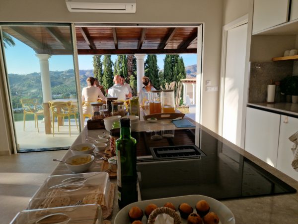 A group of six people has breakfast outside, while sun shines, picture is made from the kitchen table inside