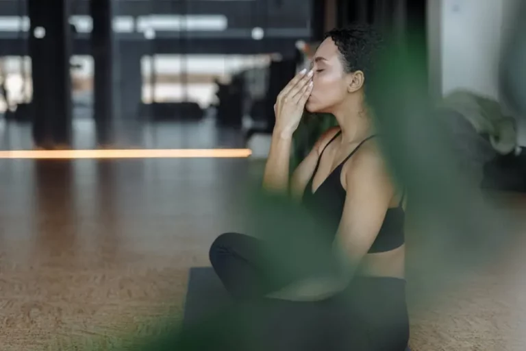 A woman in athletic wear sits cross-legged on a yoga mat indoors, practicing pranayama breathing with her eyes closed, touching her face with one hand. A green plant is partially visible in the foreground.