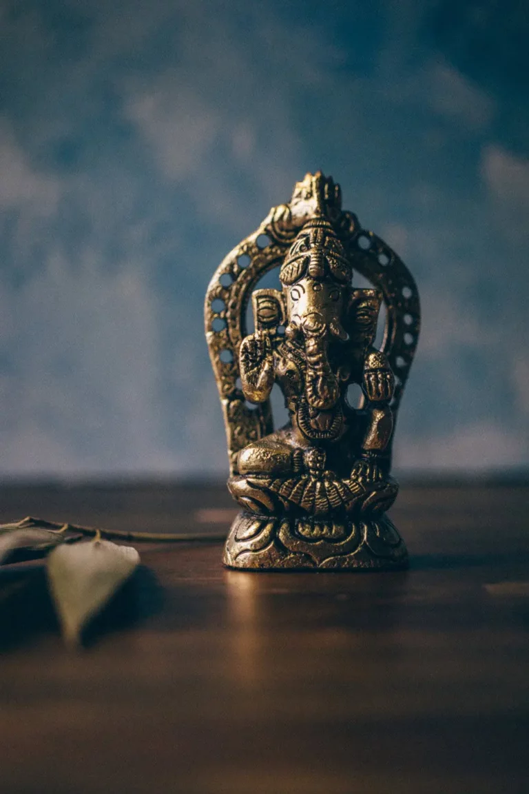 A brass statue of the Hindu deity Ganesha sits on a wooden surface with leaves beside it, set against a blurred blue background.