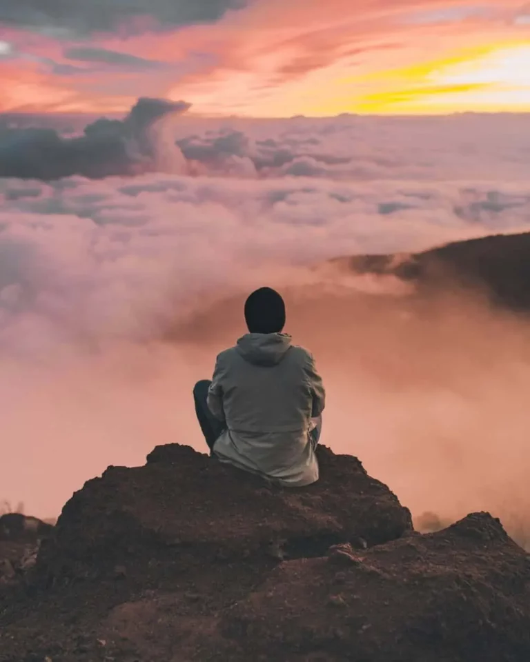 A person in a jacket and beanie sits on a rocky cliff, facing a sea of clouds and a colorful sunset sky, creating a peaceful and contemplative scene.