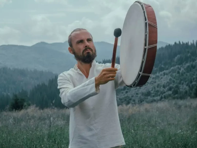 A man with a beard, wearing a white shirt, stands outdoors in a grassy field with mountains and trees in the background, playing a large drum with a mallet.