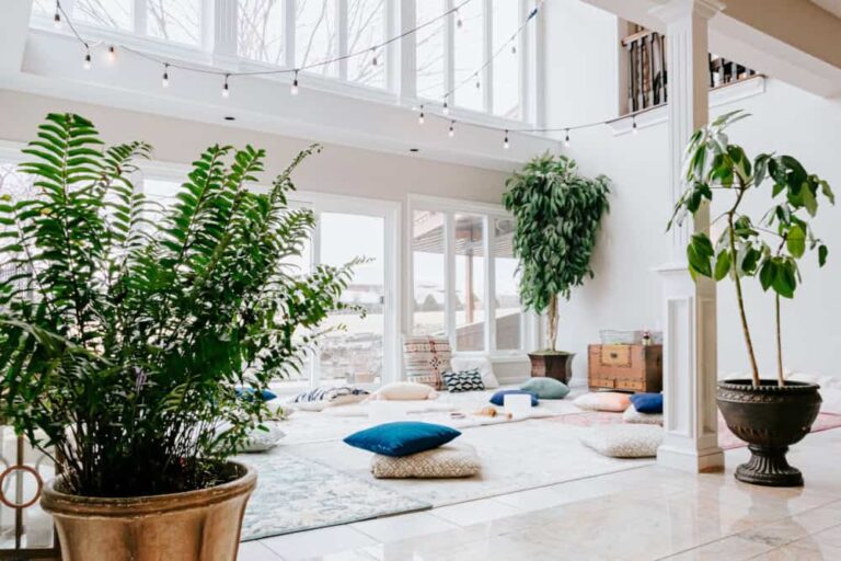 A bright, airy room with large windows, string lights, and potted plants. Floor cushions, pillows, and rugs are arranged for comfortable seating, creating a relaxing, inviting atmosphere.