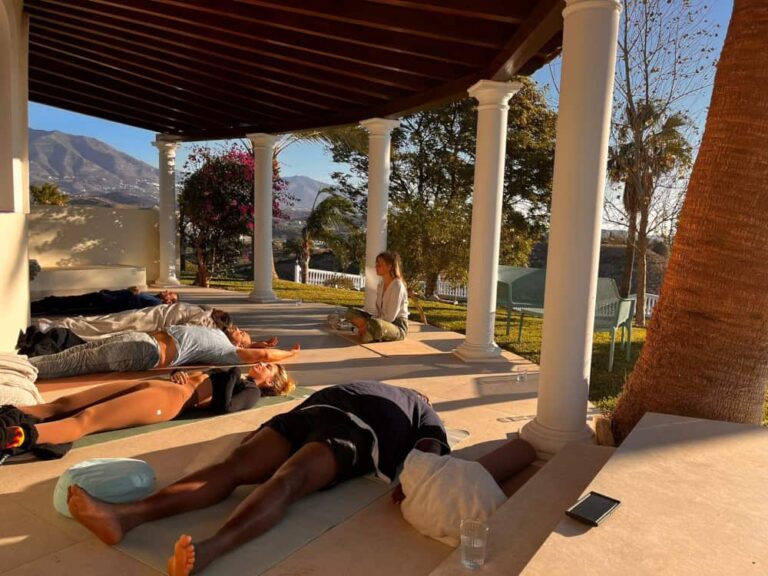 A group of people practice yoga, lying on mats in a shaded patio with columns. A woman sits cross-legged, leading the session. Sunlight streams in, and there are trees, mountains, and a glass of water nearby.