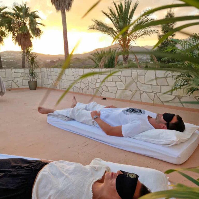 Zwei Personen liegen mit Augenmasken auf Matten im Freien und entspannen sich bei Sonnenuntergang. Palmen und Steinmauern umgeben sie, im Hintergrund sind Berge zu sehen, die eine friedliche, ruhige Atmosphäre schaffen.