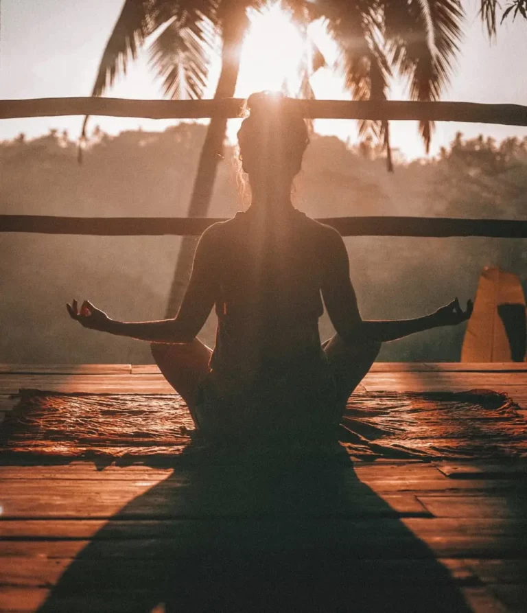 A person sits cross-legged on a mat, meditating at sunrise or sunset on a wooden deck with palm trees in the background, sunlight streaming through their silhouette.