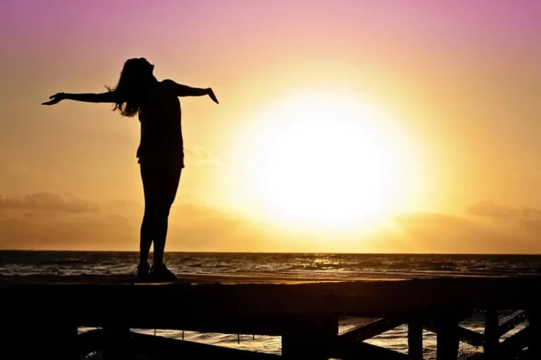 Silhouette of a person standing on a pier with arms outstretched, facing a bright sunset over the ocean, under a colorful sky.