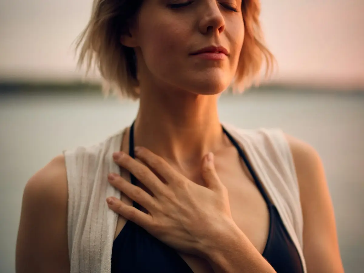 A woman with short blonde hair stands outdoors with her eyes closed, gently touching her chest with one hand. She appears calm and peaceful, wearing a sleeveless top and a light shawl.