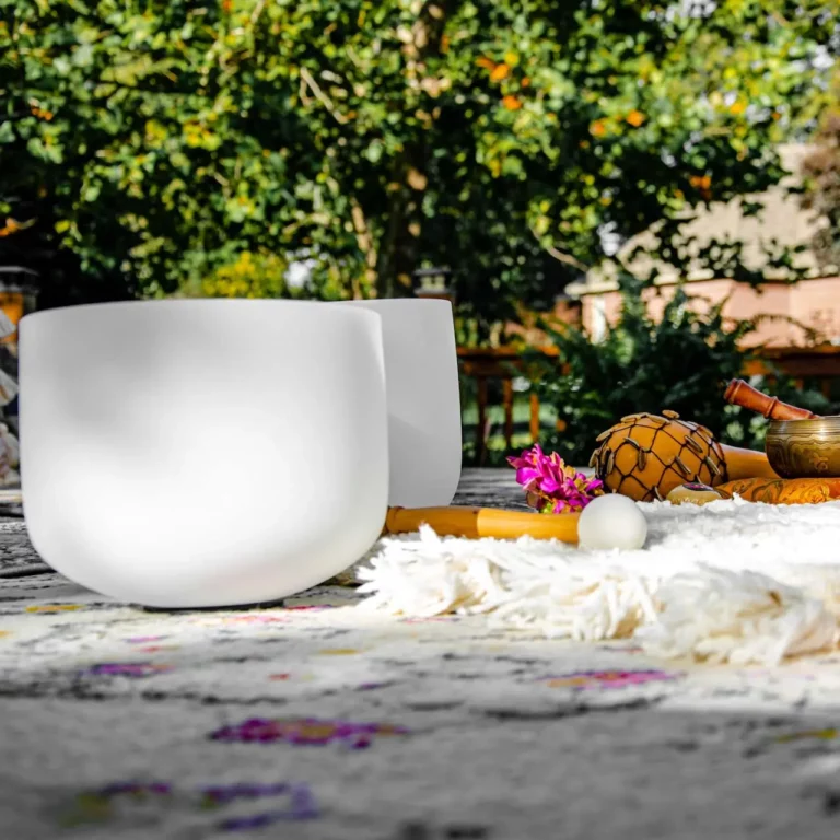 A white singing bowl sits outdoors on a patterned rug with another bowl, a mallet, small percussion instruments, and pink flowers. Green trees and a wooden fence are visible in the sunlit background.