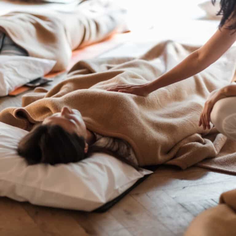 Woman laying on white mattress with brown blanket, another woman touching her chest