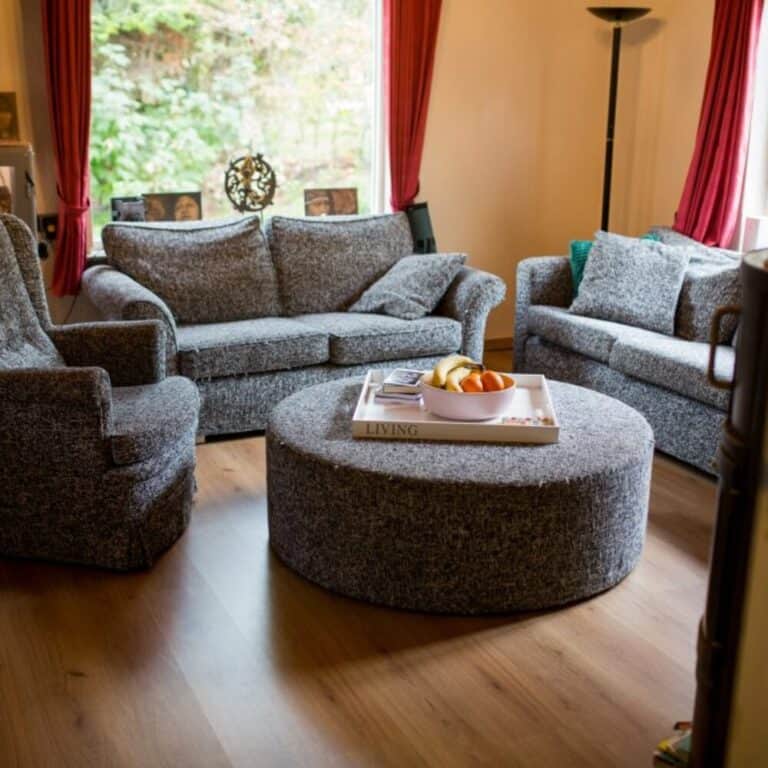 A cozy living room with two gray sofas, a gray armchair, and a large round gray ottoman. The ottoman has a white tray with a bowl of fruit and books on top. The room has wooden floors and a large window with red curtains letting in natural light.