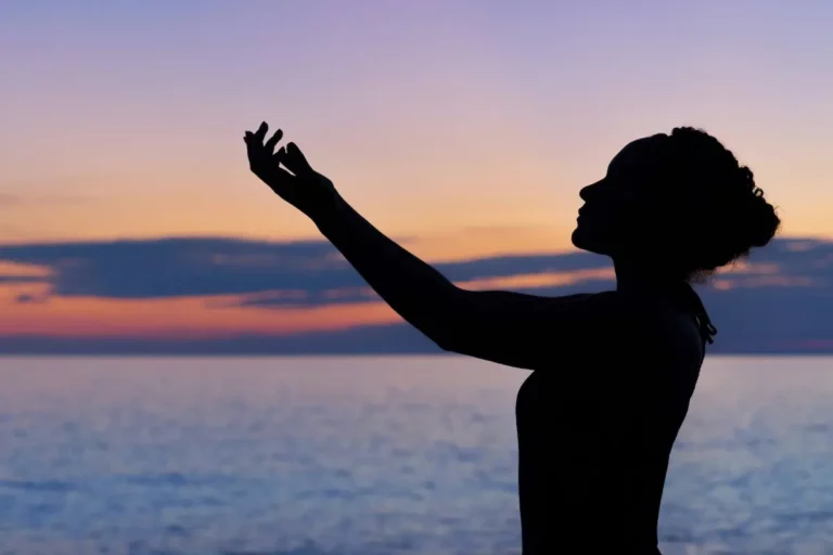 Silhouette of a person with arms raised, standing by the sea during a colorful sunset with orange, pink, and blue hues in the sky.