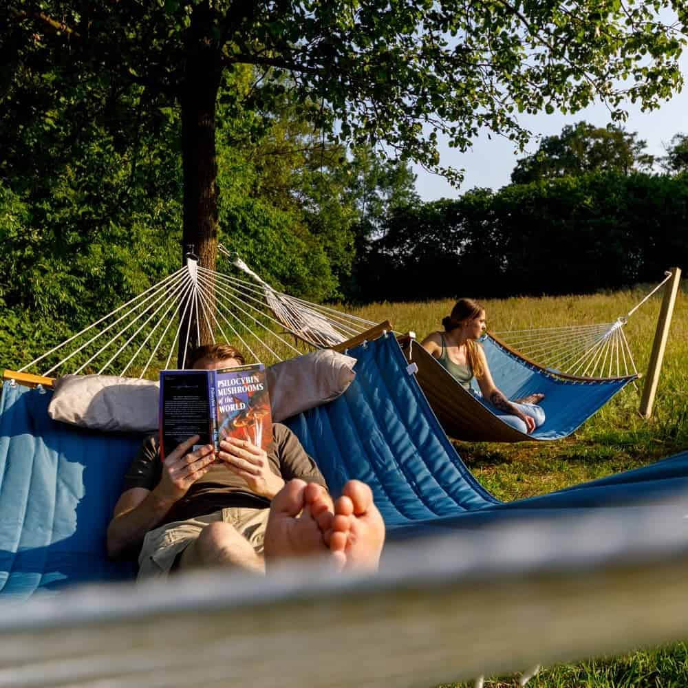 Zwei Personen entspannen sich in blauen Hängematten unter einem Baum in einem grasbewachsenen Feld. Die Person im Vordergrund liest ein Buch mit dem Titel 