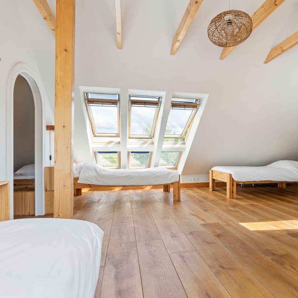 A bright attic bedroom with wooden flooring, three beds, and skylight windows letting in natural light. The room features exposed wooden beams.