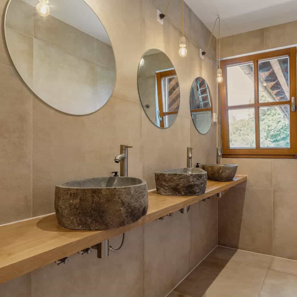 A modern bathroom features stone sinks, round mirrors, hanging light bulbs, and a wooden window overlooking a green outdoor area. Neutral tone tiles adorn walls.