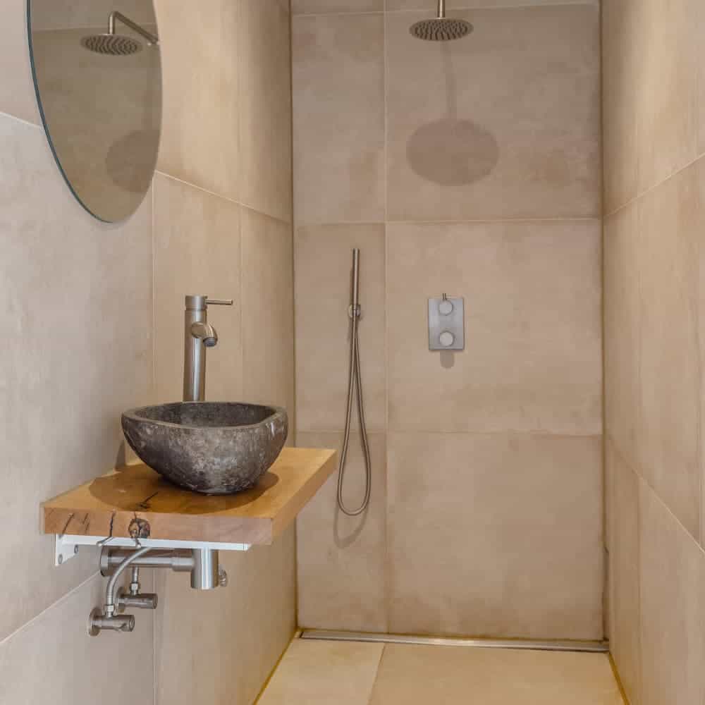 Modern bathroom with a walk-in shower, round mirror, stone basin sink on a wooden shelf, and beige tiled walls. Minimalist design with overhead shower.