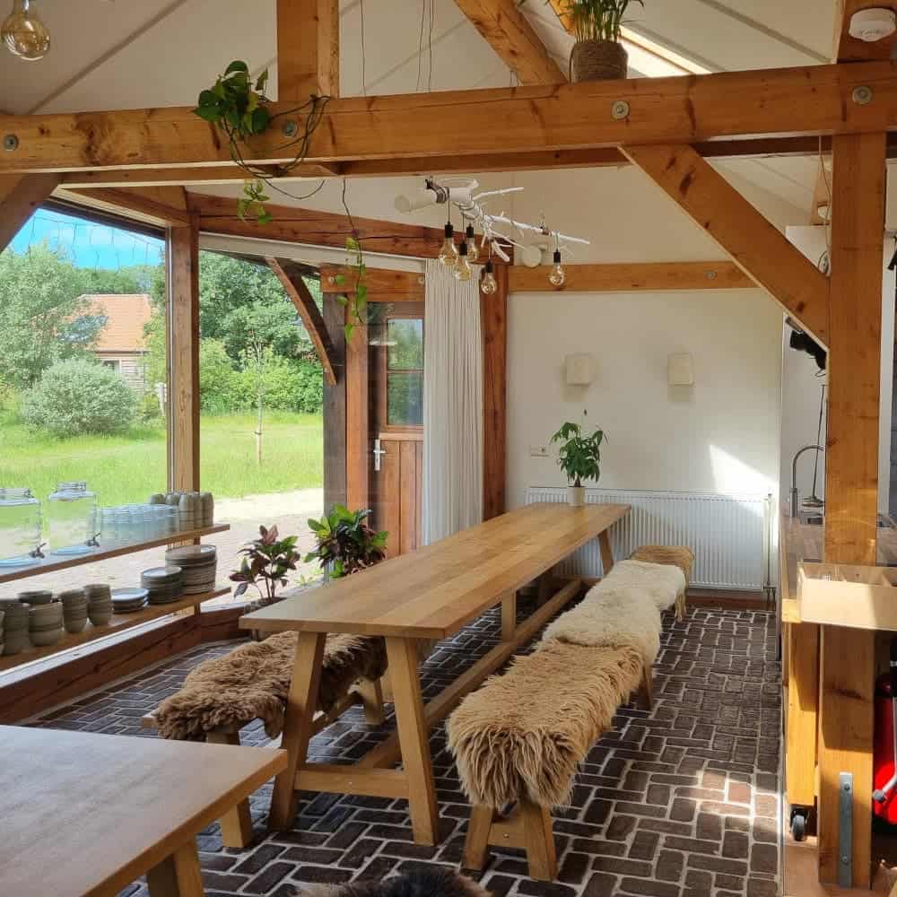 A cozy, rustic wooden dining area with large windows overlooking a green yard, featuring long tables, sheepskin-covered benches, and potted plants.