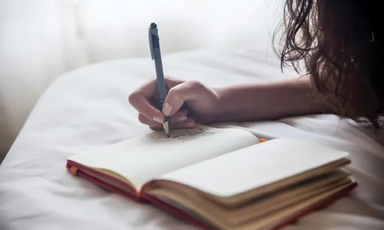 A person with long hair is lying on a bed and writing in an open notebook with a pen, near a softly lit window.