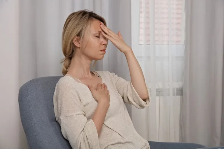 A woman sits on a chair indoors, eyes closed, with one hand on her forehead and the other on her chest, appearing stressed or unwell. Light curtains and a window are in the background.