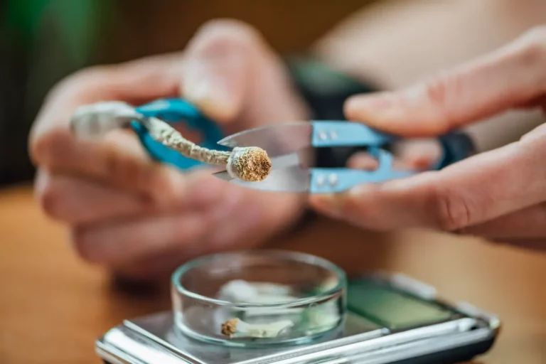 Hands use scissors to cut dried psilocybin mushrooms over a glass dish on a digital scale, suggesting careful weighing or preparation of the mushrooms.