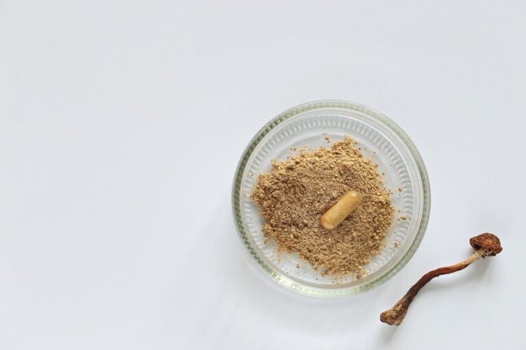 A glass bowl containing ground psilocybin mushrooms powder with a capsule on top, placed next to a dried mushroom on a white background.