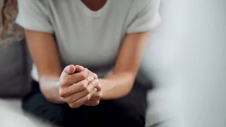 A person sits on a couch with hands clasped together, leaning slightly forward. Their face is out of frame, and they wear a plain white t-shirt and dark pants. The mood appears contemplative or anxious.
