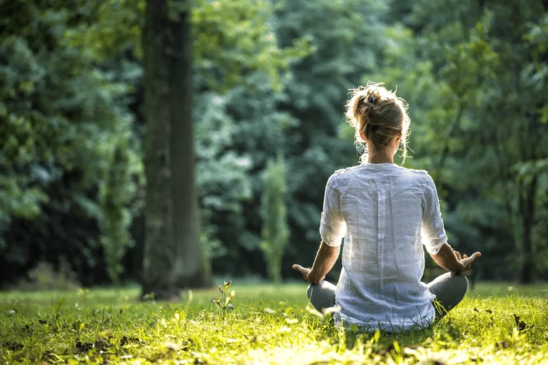 A person sits cross-legged on grass in a sunlit forest, meditating with hands resting on knees. They wear a white shirt and face away from the camera, surrounded by green trees and natural light.