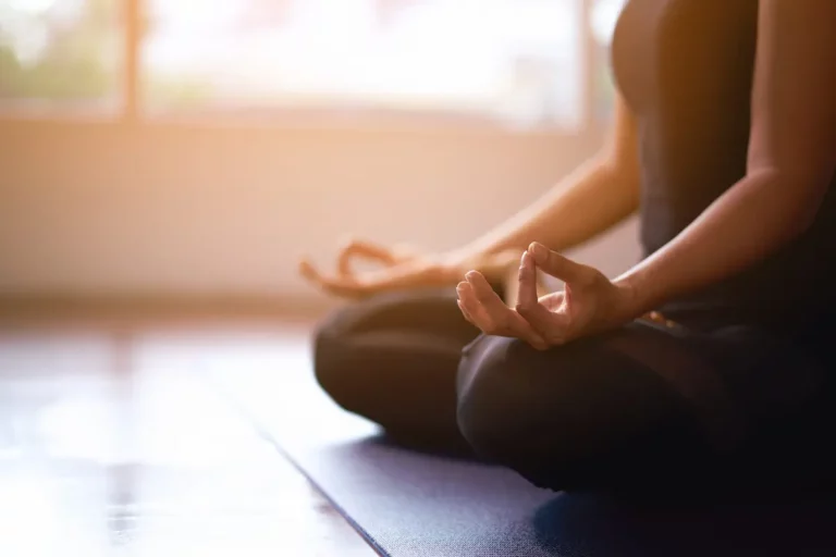 A person sits cross-legged on a yoga mat indoors, with hands resting on knees in a meditation pose, fingers forming a mudra. Sunlight streams through a window, creating a calm and peaceful atmosphere.
