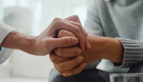 Close-up of one person gently holding another person's hands, offering comfort and support. Both individuals are wearing long sleeves, and the scene conveys empathy and compassion.