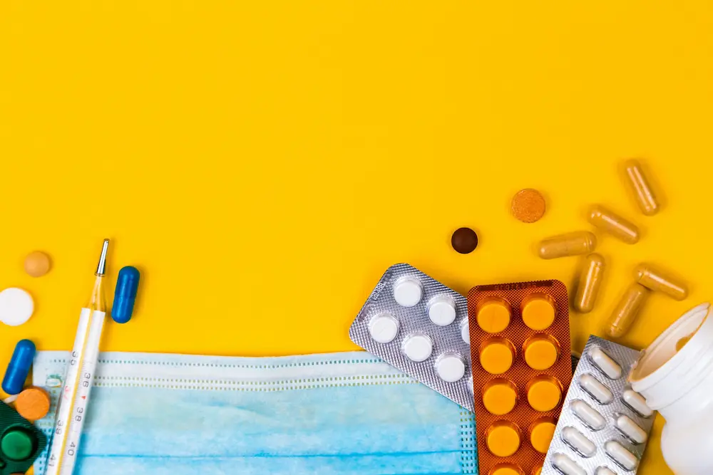 Assorted pills, tablets, and capsules, a face mask, a thermometer, and a pill bottle are arranged on a bright yellow background with empty space at the top.
