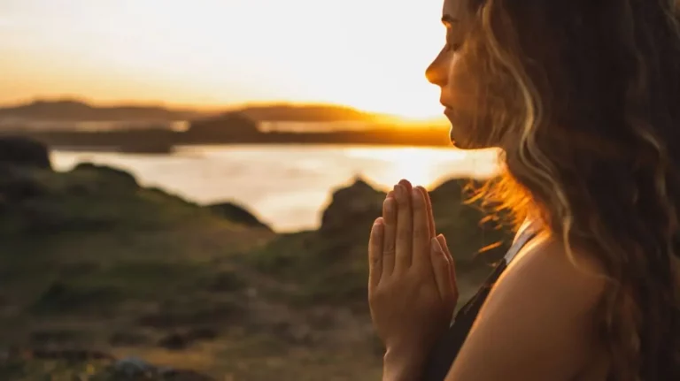 A woman with long hair is standing outdoors at sunset, with her hands pressed together in a prayer position, facing the light near a body of water and grassy landscape.