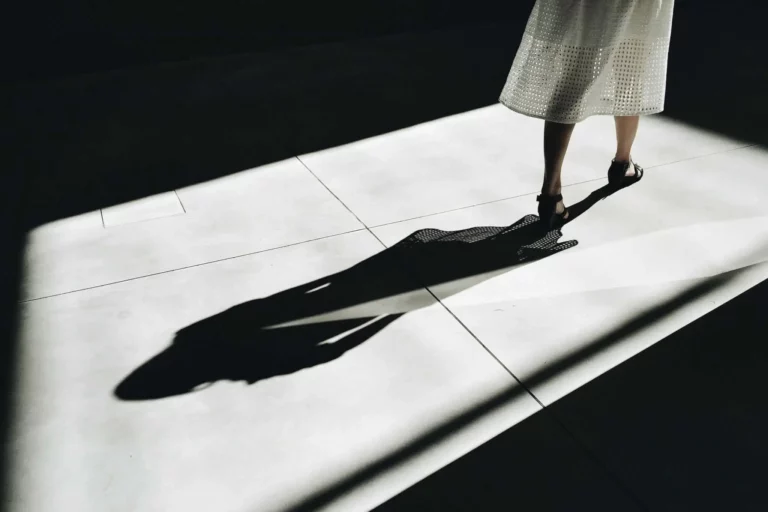 A person wearing a white knee-length skirt and black shoes stands in a beam of sunlight, casting a long shadow on a tiled floor surrounded by darkness.