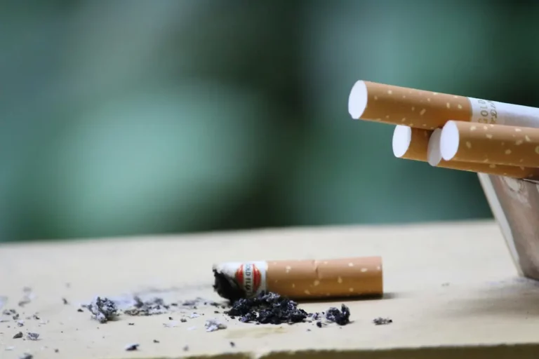Close-up of a broken cigarette lying on a surface next to ashes, with a pack of unlit cigarettes partially visible in the background. The scene is set against a blurred green backdrop.
