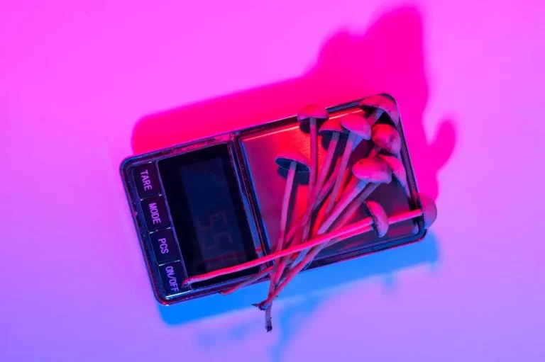 A digital scale with several psilocybin mushrooms on top, illuminated by pink and purple lighting, sits on a white surface.