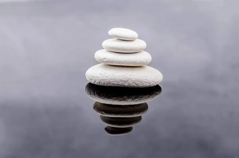 Four smooth white stones are stacked in a pyramid shape on a reflective surface, creating a clear mirror image beneath them. The background is blurred and gray, emphasizing calm and balance.