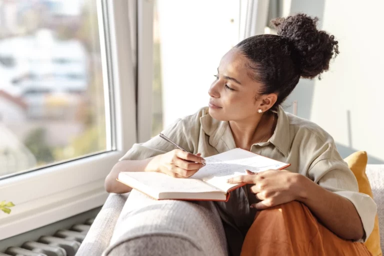 A woman sits on a couch by a large window, holding an open notebook and pen. She looks thoughtfully outside, appearing to be in deep thought or reflection as she writes.