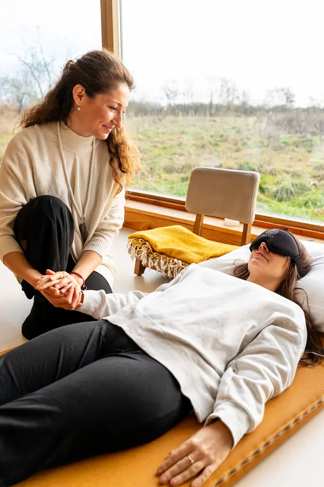 A woman sits beside another woman lying on a mat, holding her hand during a psilocybin session. The woman lying down wears an eye mask, while the other smiles supportively. Large windows reveal a grassy landscape outside.