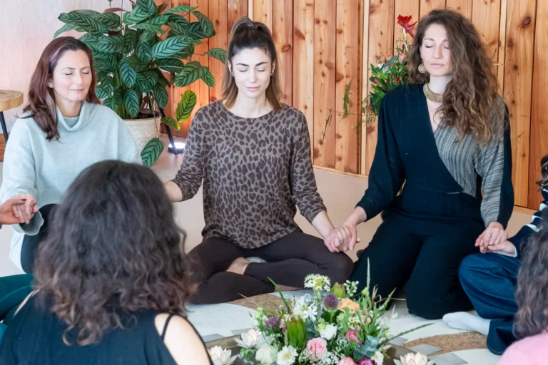 Four women sit cross-legged in a cozy, wood-paneled room, surrounded by plants and flowers, holding hands as they meditate together with eyes closed.