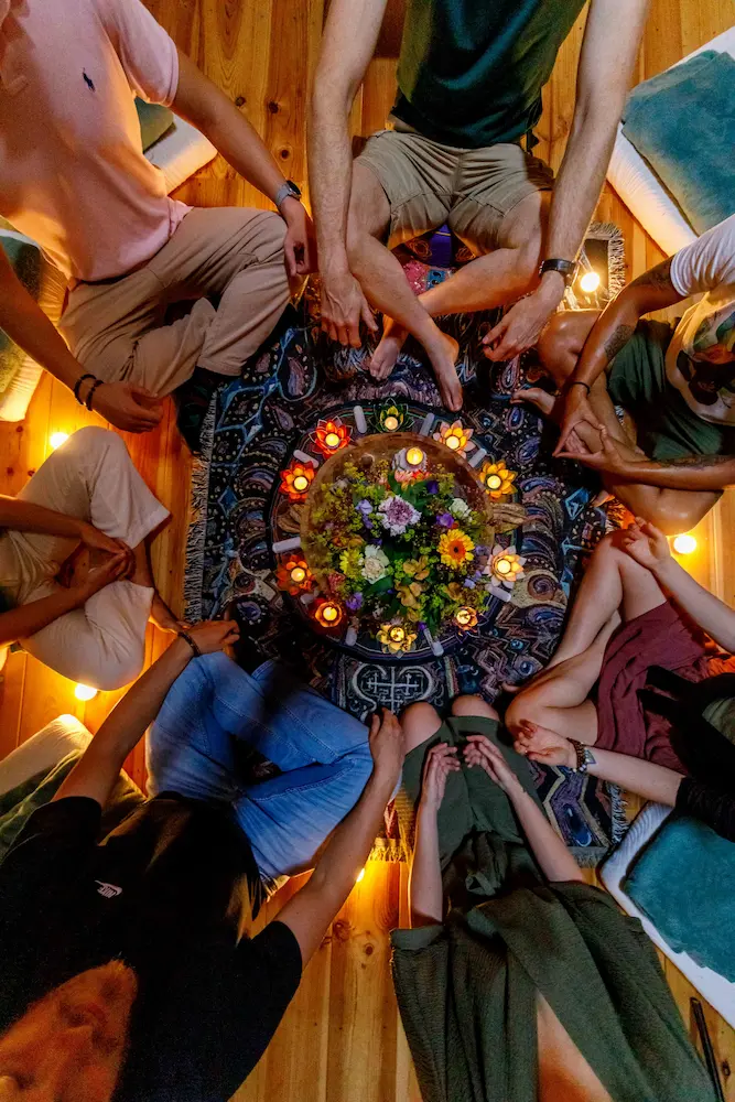 A group of people sit cross-legged in a circle around a centerpiece of flowers and candles on a patterned rug, in a warmly lit wooden room, viewed from above.