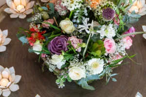 A colorful floral arrangement with roses, gerbera daisies, and greenery sits on a round table, surrounded by lit lotus-shaped candle holders.