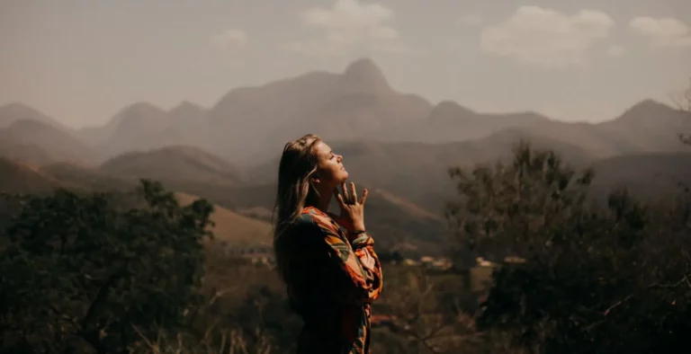 A woman stands outdoors with her eyes closed and head tilted up, hands near her face in a serene gesture, with mountains and a hazy sky in the background.