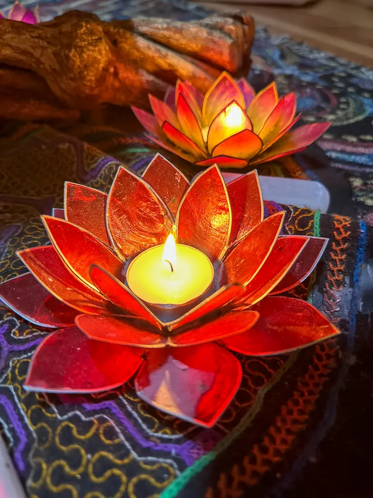 A lit tealight candle sits inside a vibrant red lotus-shaped holder, with another similar holder in the background, both placed on a colorful patterned surface.