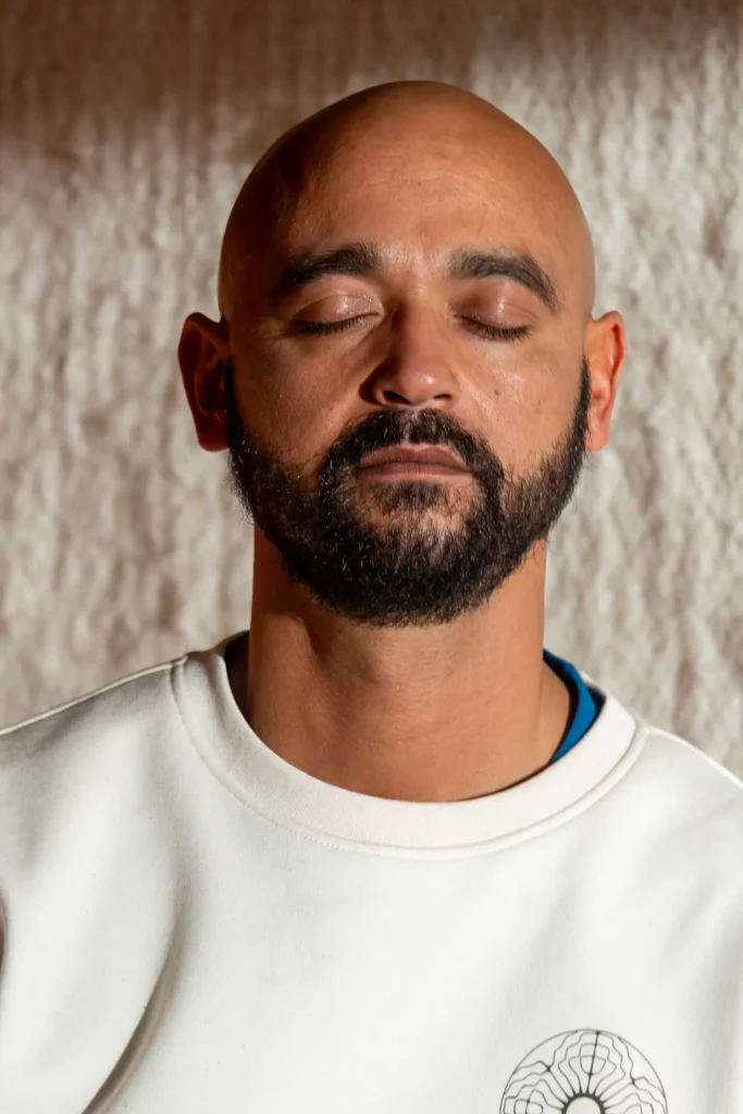 A man with a bald head and beard wearing a white sweatshirt sits with his eyes closed, appearing calm and relaxed as he meditates against a light textured background.