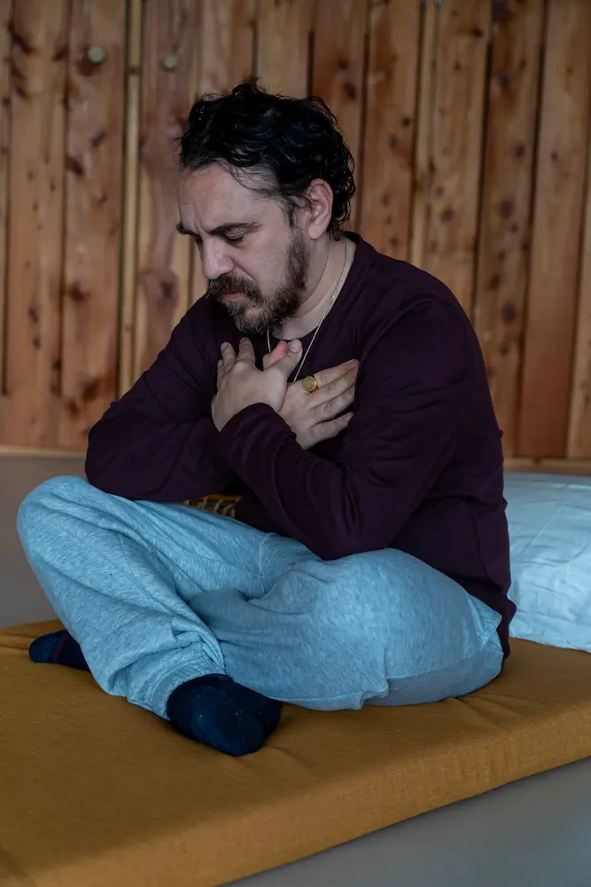 A man with dark hair and a beard sits cross-legged on a mat, eyes closed and hands crossed over his chest, appearing calm as he meditates. Wooden wall panels are in the background.