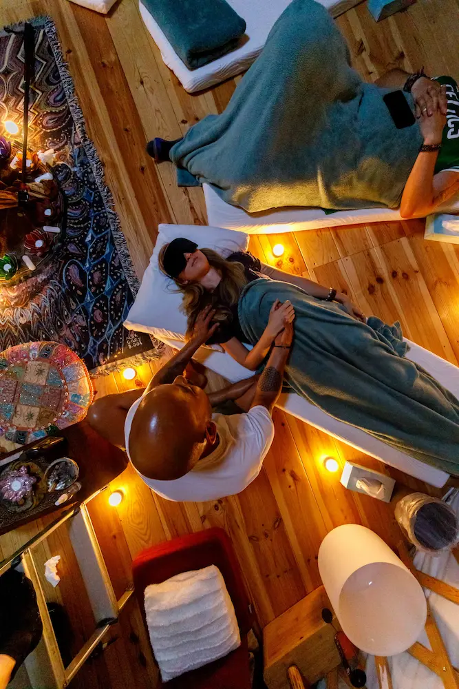 A person lies on a mat covered with a green blanket, eyes closed, while another gently places hands on their chest during a psilocybin ceremony in a dim, candle-lit room with a cozy, relaxed atmosphere.