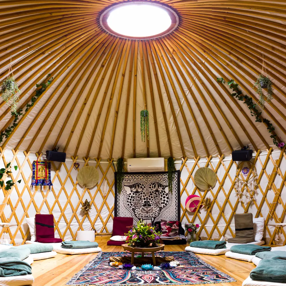 Interior of a cozy yurt with a circular skylight, wooden beams, cushions in a circle on the floor, colorful rugs, plants, and decorative wall hangings, creating the warm, inviting atmosphere typical of a traditional yurt.