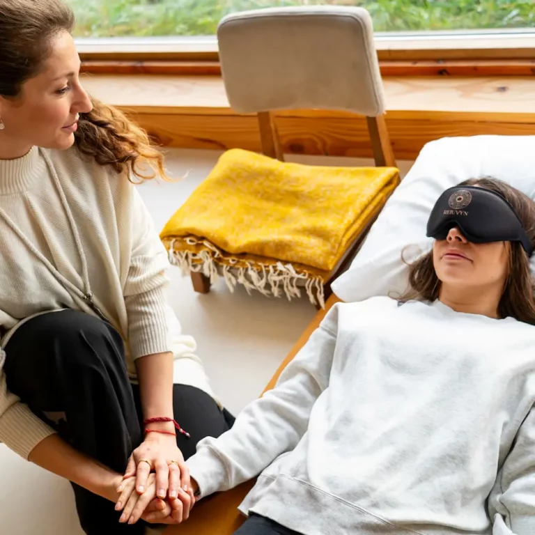 A facilitator in a white sweater and black pants sits on the floor, holding hands with a customer who lies on a mat during a serene psilocybin ceremony. The person lying wears a gray sweatshirt and an eye mask, surrounded by blankets and pillows, with a soothing grassy view outside.
