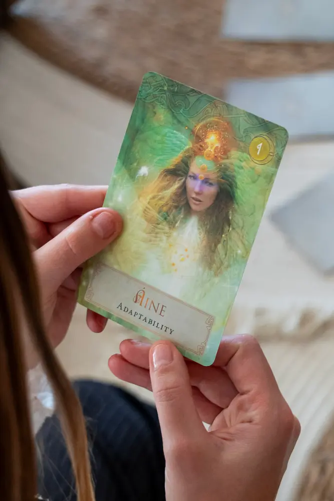 A person holds a tarot card featuring a mystical woman with flowing hair and glowing symbols, labeled "Aine" and "Adaptability." The background is green and ethereal.