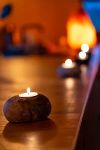 A small tealight candle sits in a smooth stone holder on a wooden surface, with more lit candles blurred in the background, creating a warm and calming atmosphere.