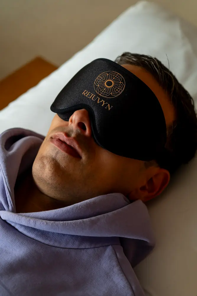 A person wearing a black eye mask with the word "REJUVYN" and a gold design, lying on a pillow in a light purple hoodie, evokes calm during a psilocybin session.