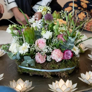 A floral arrangement with pink roses, white and yellow flowers, and greenery sits on moss in the center of a round tray, surrounded by lotus-shaped candle holders. People are seated around the display.